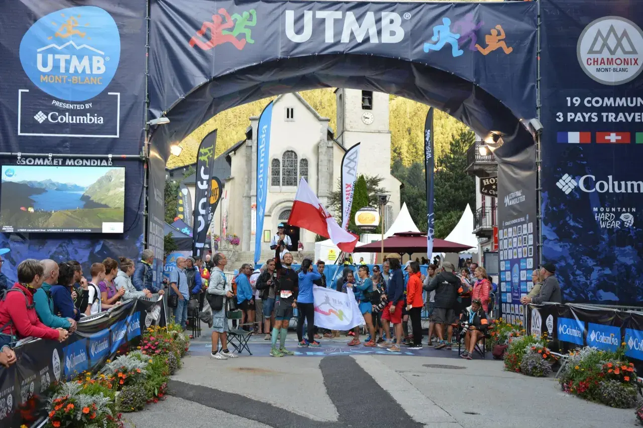 Uczestnicy Ultra-Trail du Mont-Blanc świętują pod łukiem startowym. W tle kościół i tłum widzów.