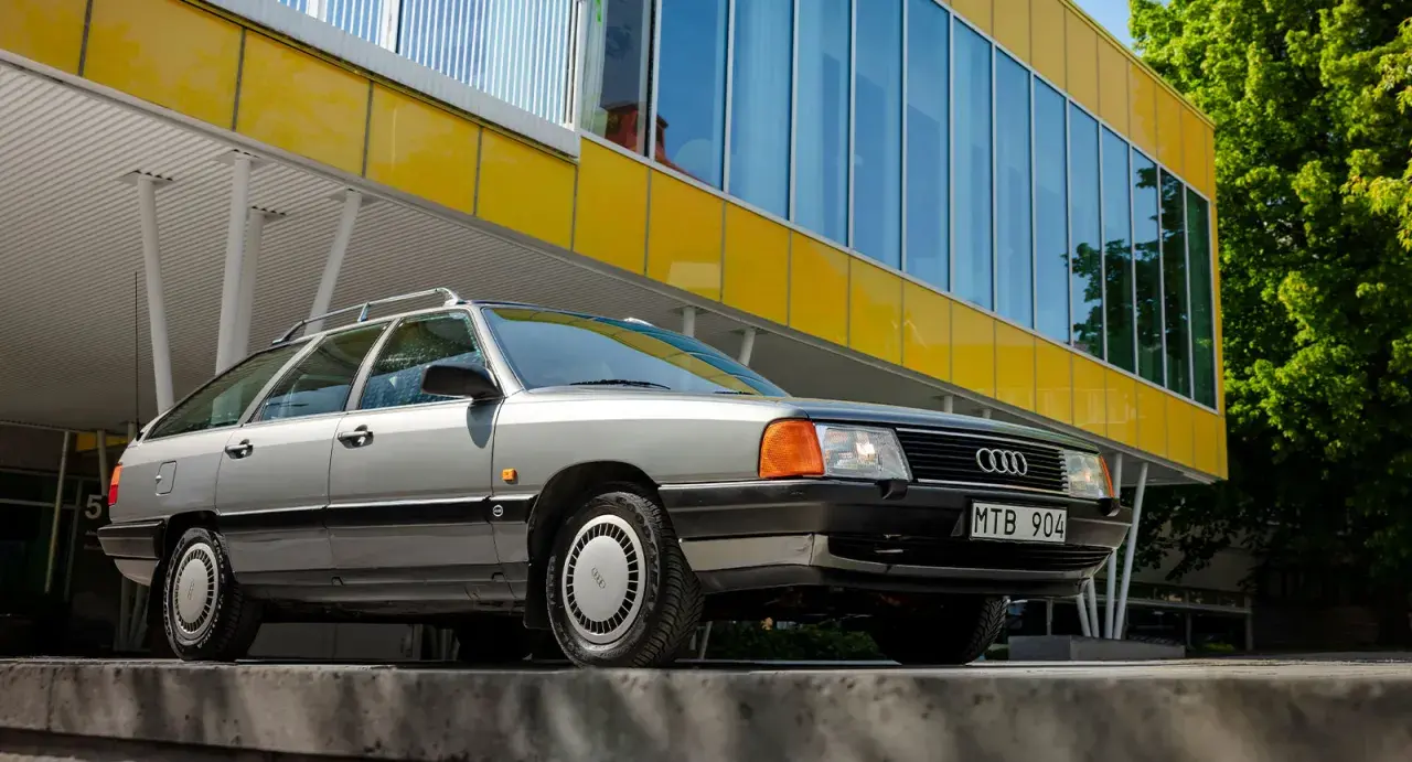 En grå Audi 100 C3 Avant står parkerad framför en modern gul byggnad med stora fönster.
