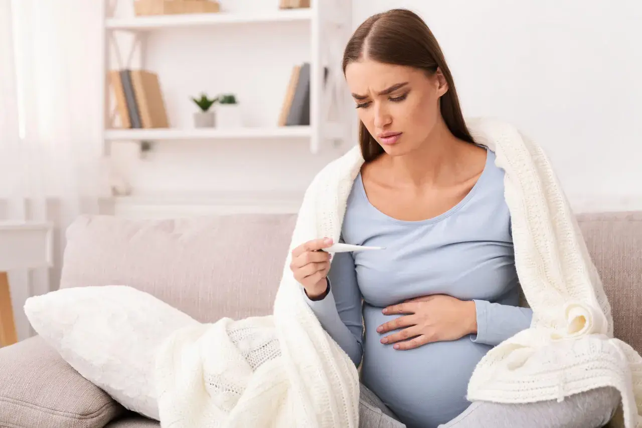 Donna incinta preoccupata guarda il termometro, avvolta in una coperta. La febbre in gravidanza è un sintomo da non sottovalutare.