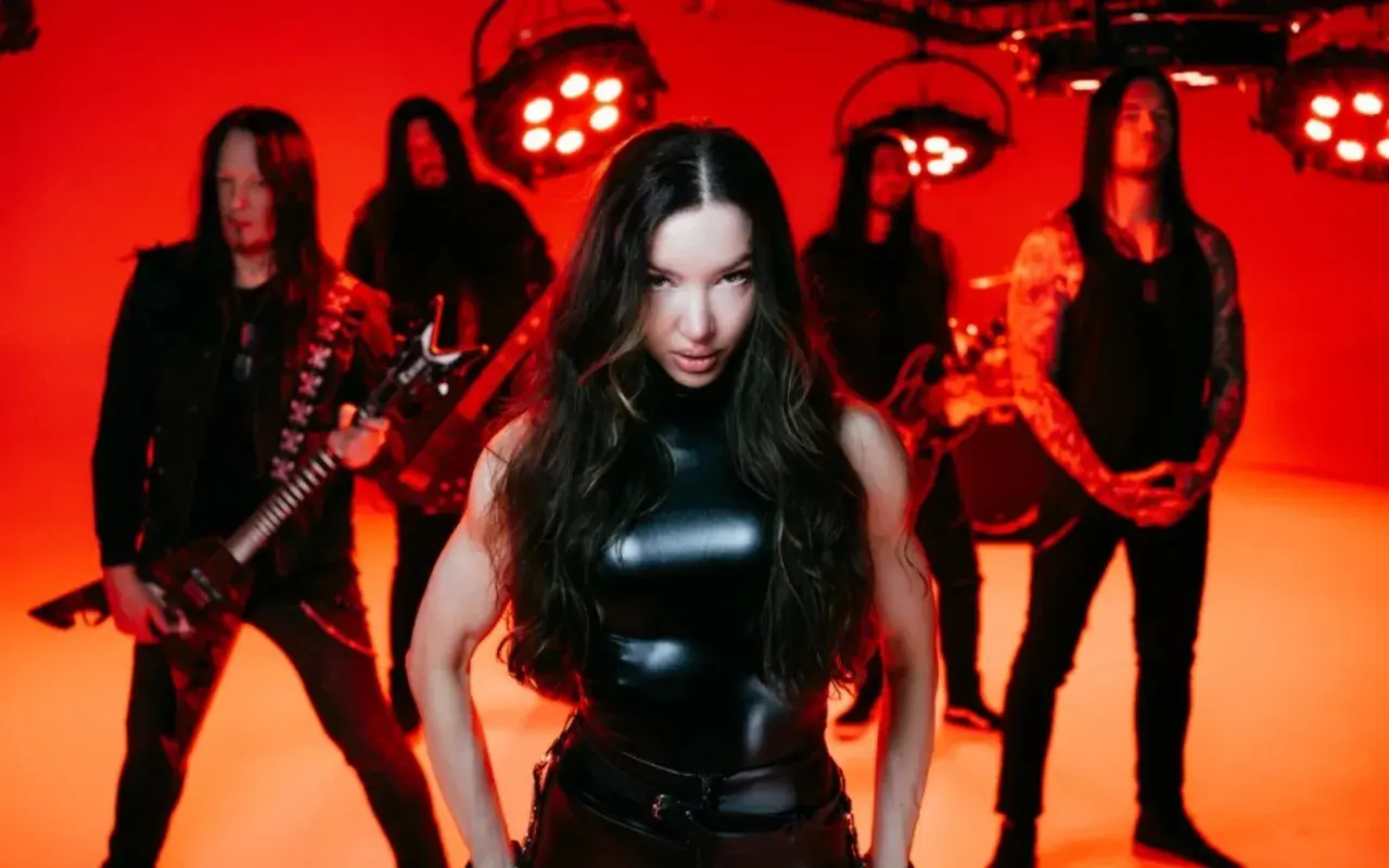 Arch enemy wokalistka w skórzanym stroju, otoczona przez zespół na tle czerwonych świateł.