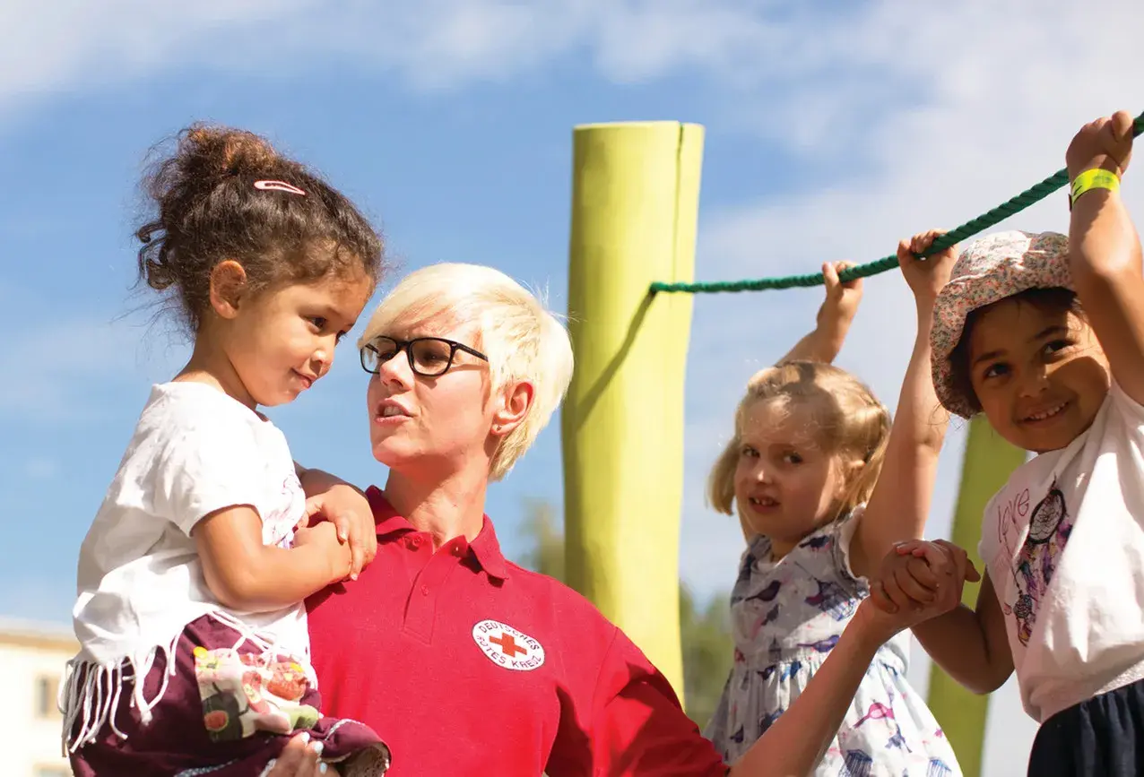Frau vom Roten Kreuz fördert **Inklusion im Kindergarten**, indem sie ein Kind hält, während andere Kinder an einem Seil spielen.