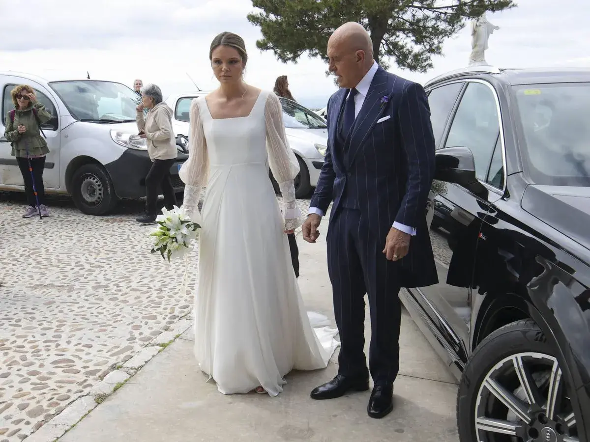 Novia con ramo de lirios y su acompañante, un hombre con traje de raya diplomática, llegan a su boda. Irene Matamoros, radiante.