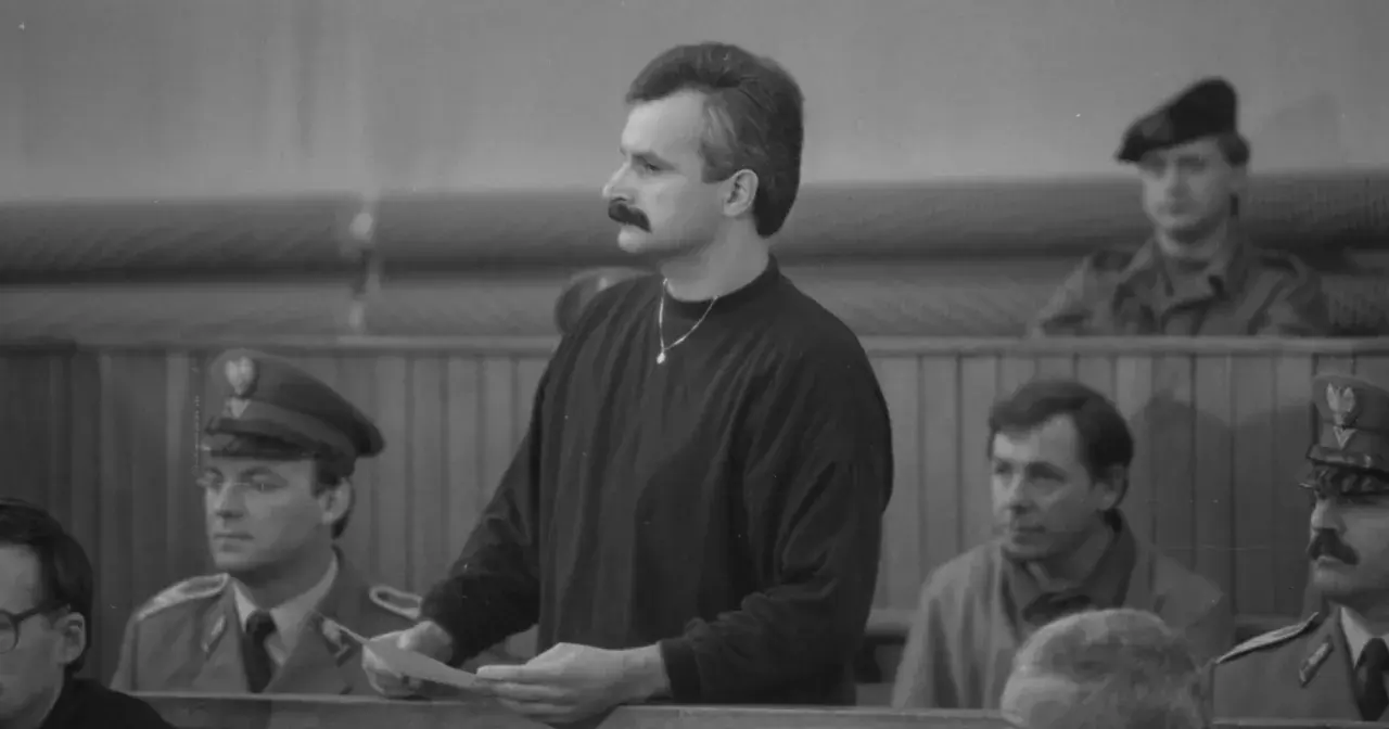 Mężczyzna z wąsami, niczym minstrel, trzyma dokumenty w sali sądowej. Obok niego siedzą ludzie w mundurach.