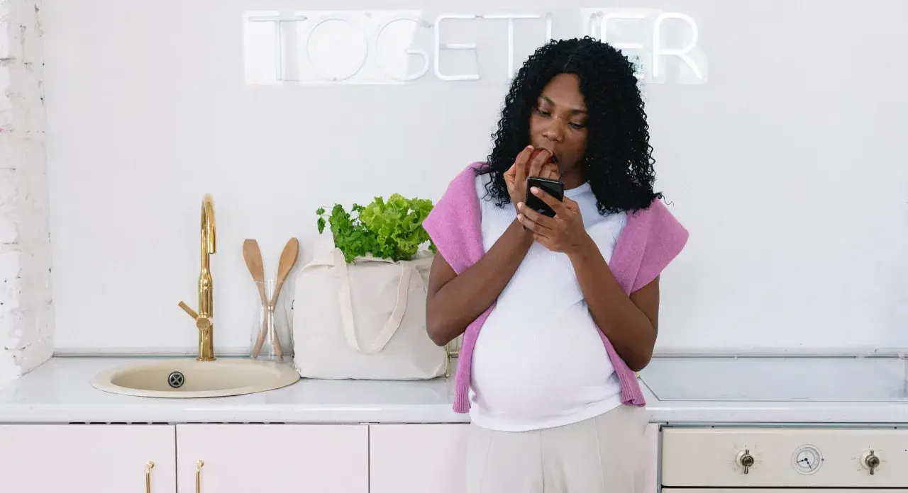 Gravid kvinna i köket kollar mobilen och äter ett äpple, en påminnelse om vad man ska undvika som gravid.