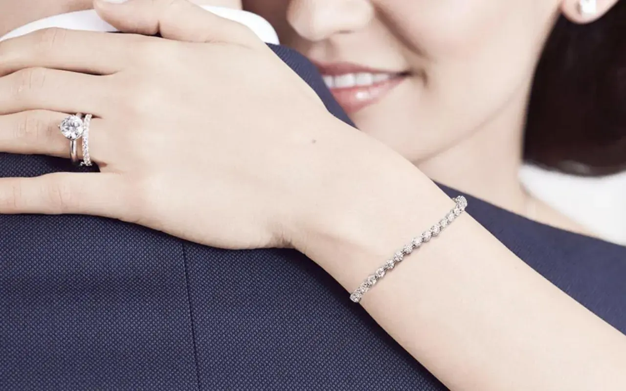 Anillo de compromiso en la mano izquierda, símbolo de amor eterno. Una mujer abraza a su pareja, luciendo un brazalete de diamantes.