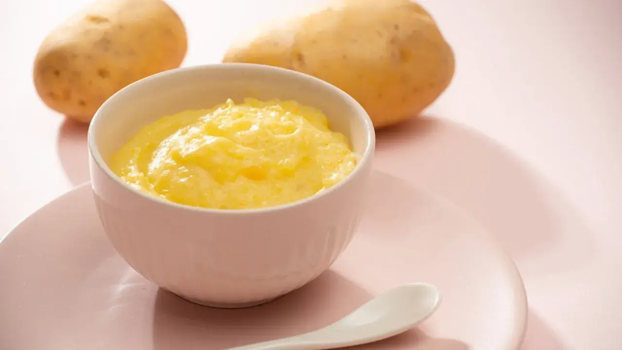 Ein Schälchen mit cremigem Kartoffelbrei, bereit für ein Baby. Zwei Kartoffeln liegen im Hintergrund.