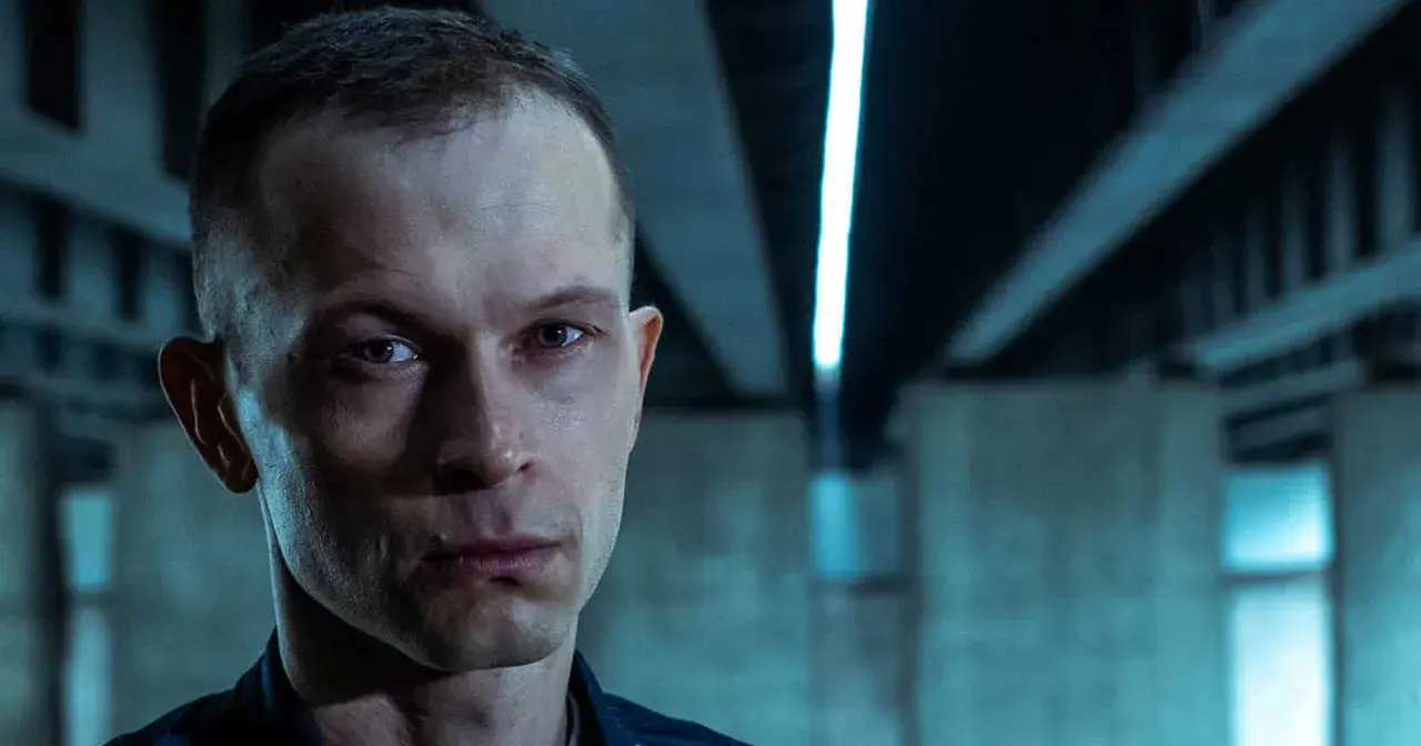 Zbliżenie na twarz mężczyzny z filmu "Tomasz Komenda". Jego spojrzenie jest intensywne, a tło sugeruje mroczne otoczenie.