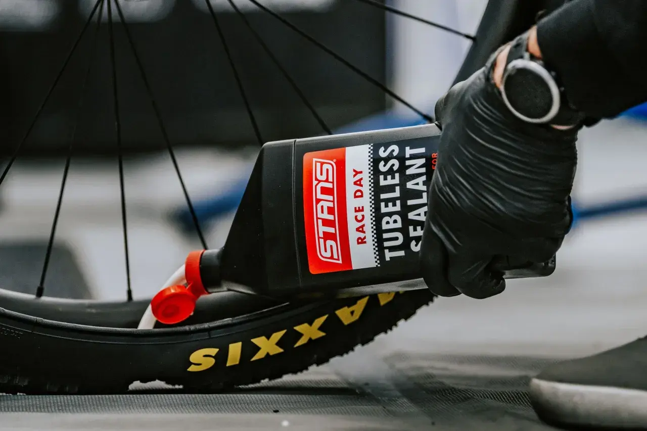 Gloved hands pour Stans Race Day tubeless sealant into a Maxxis tire, demonstrating how to add sealant to tubeless tires for a puncture-free ride.