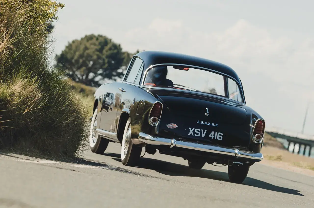 Svart Simca Aronde glider längs en kustväg, en klassisk skönhet bland gräs och träd.