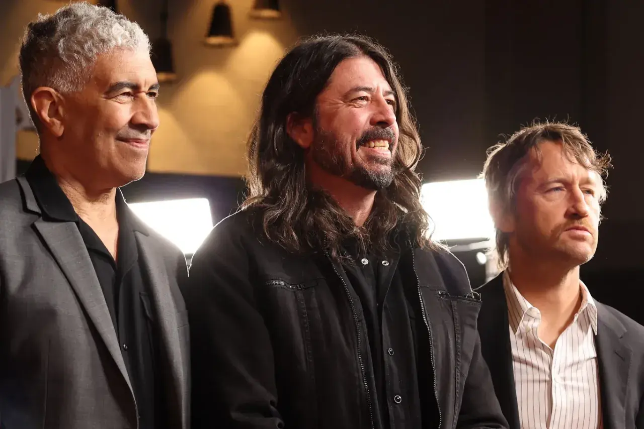 Trzech członków Foo Fighters: Nate Mendel, Dave Grohl i Chris Shiflett, uśmiechają się i patrzą w bok.