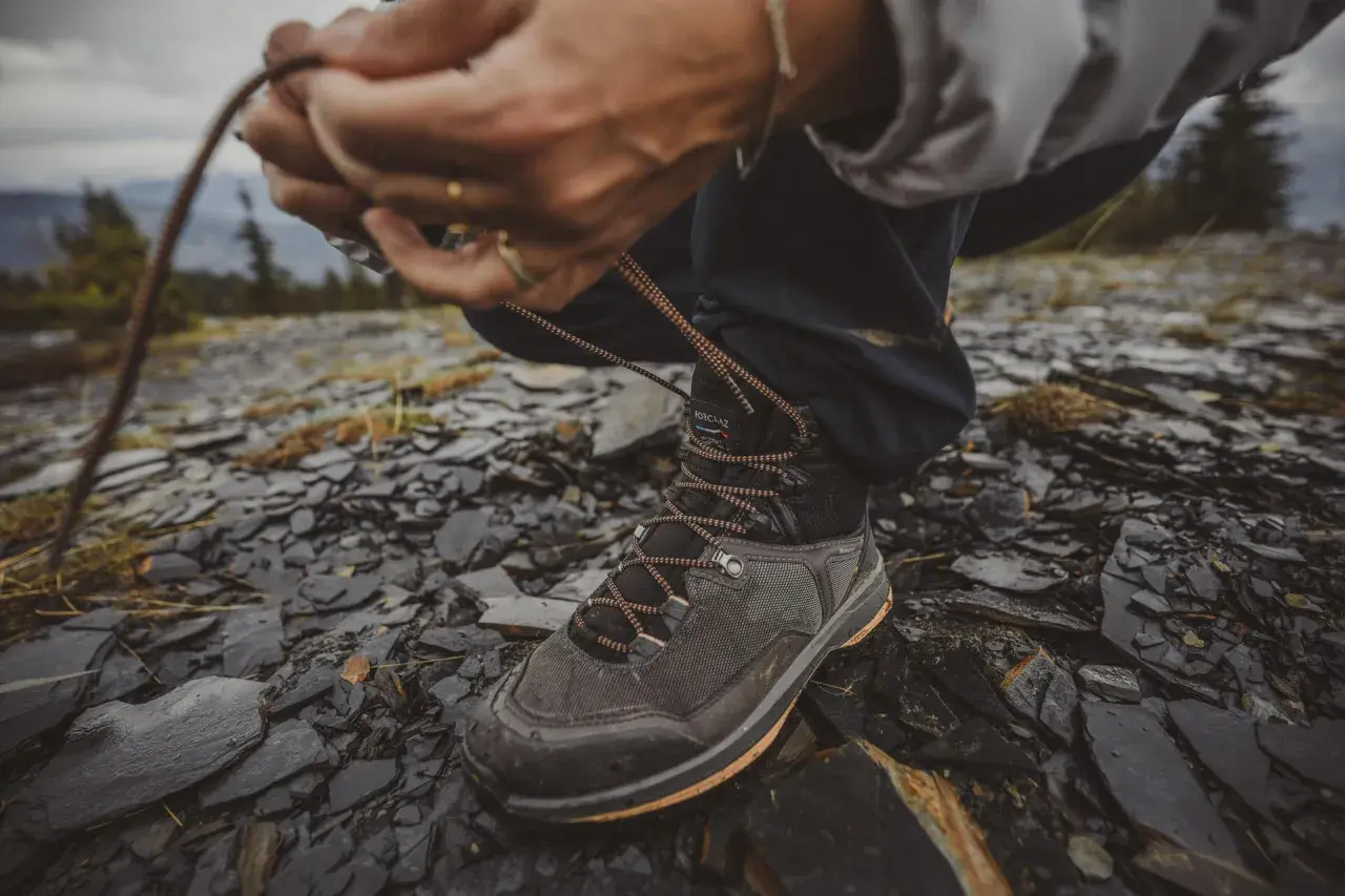 Ręce zawiązują sznurówki wysokich butów trekkingowych na kamienistym, wilgotnym podłożu.