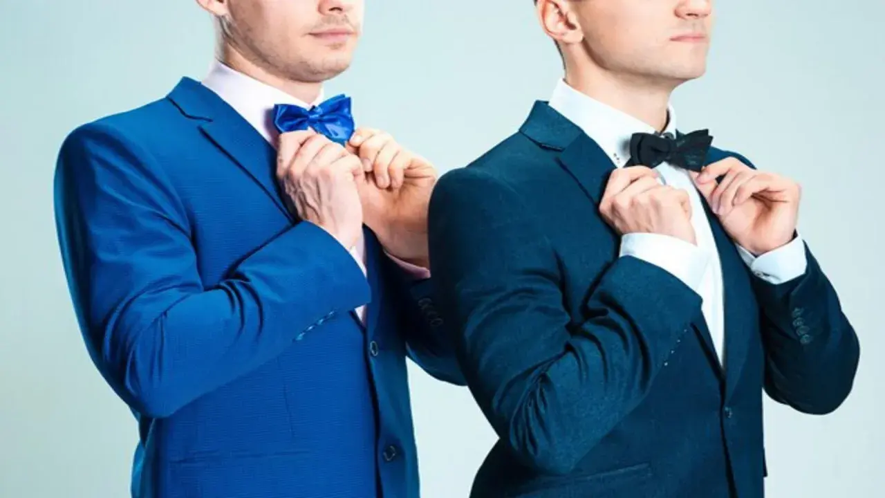 Dwóch mężczyzn w eleganckich garniturach, jeden w niebieskim, drugi w granatowym, poprawia muszki. Jaka mucha do granatowego garnituru?