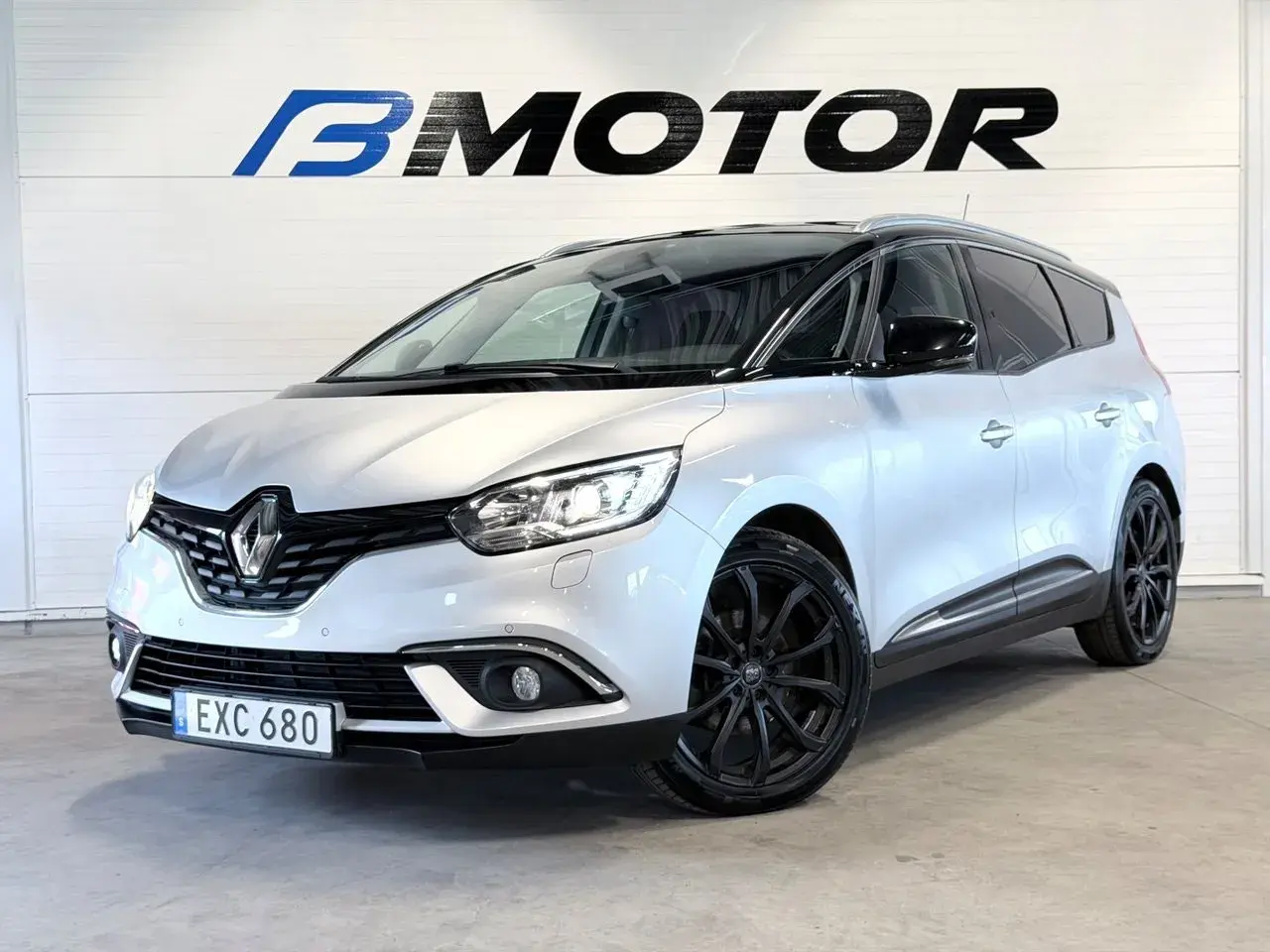 En silverfärgad Renault Grand Scénic med svarta fälgar står framför en vägg med logotypen B MOTOR.