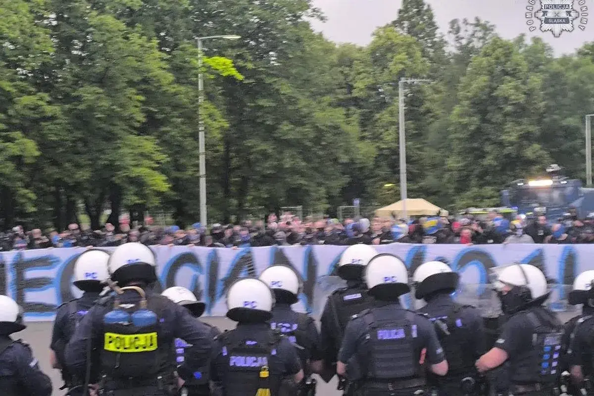 Policja w pełnym rynsztunku stoi przed grupą pseudokibiców, którzy trzymają transparent.