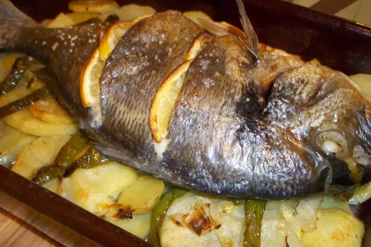 Dorada al horno con rodajas de limón, patatas y pimientos verdes en una bandeja.