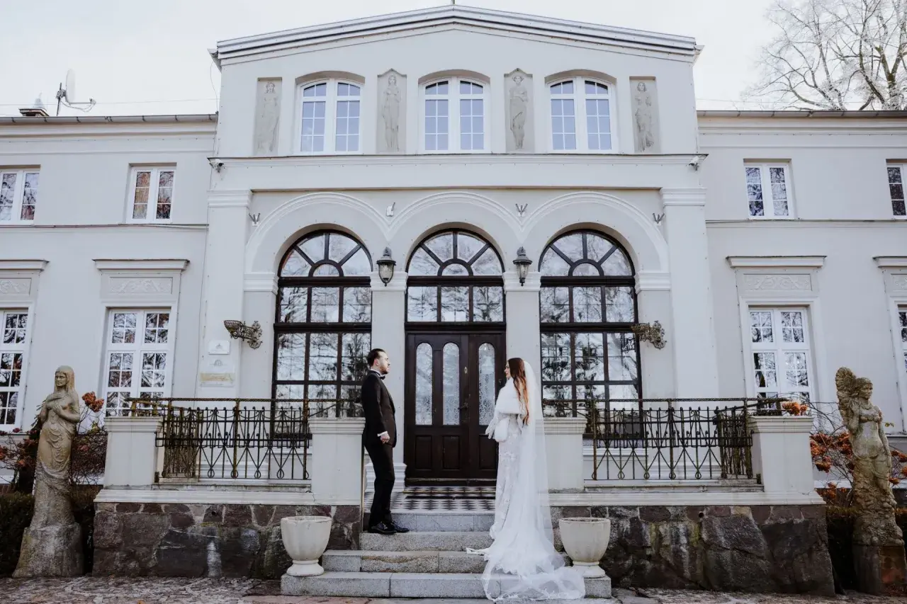 Para młoda przed Lisewskim Dworem. Elegancka biel sukni ślubnej i czarny garnitur pana młodego kontrastują z fasadą zabytkowego budynku.