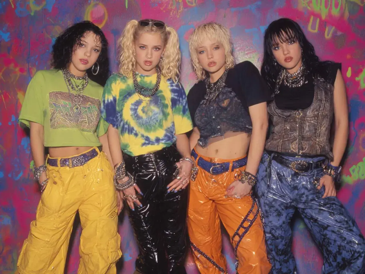 Vier junge Frauen im coolen **mode 90er jahre** Stil mit bunten Outfits und auffälligem Schmuck posieren vor einer Graffiti-Wand.