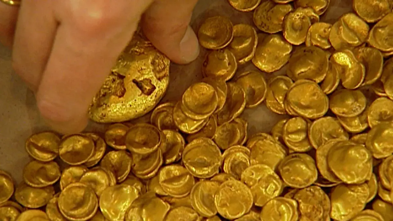 Ein Finger berührt einen Teil des Goldschatzes von Manching, der aus vielen kleinen, unregelmäßigen Goldmünzen besteht.