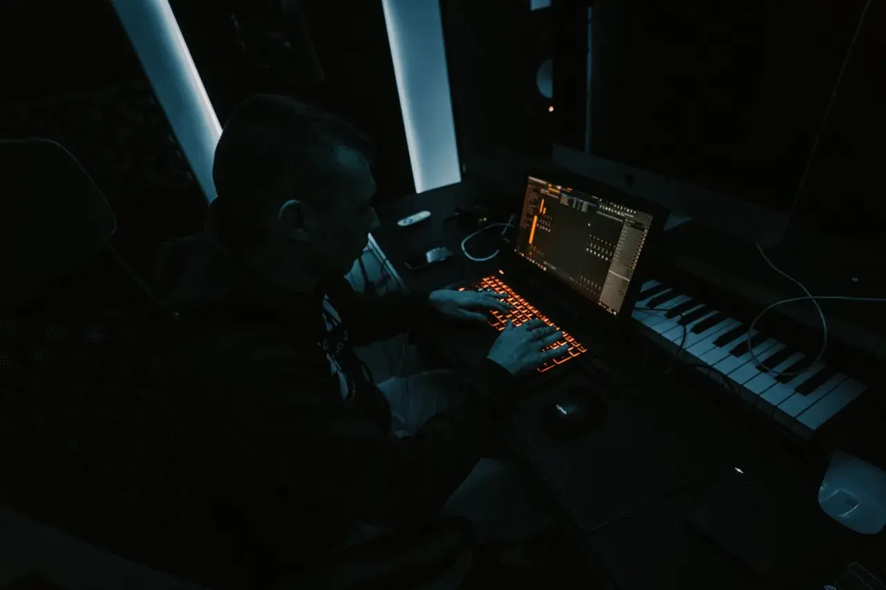 Producent muzyczny tworzy przebój w ciemnym studiu, pracując przy laptopie i klawiaturze sterującej.