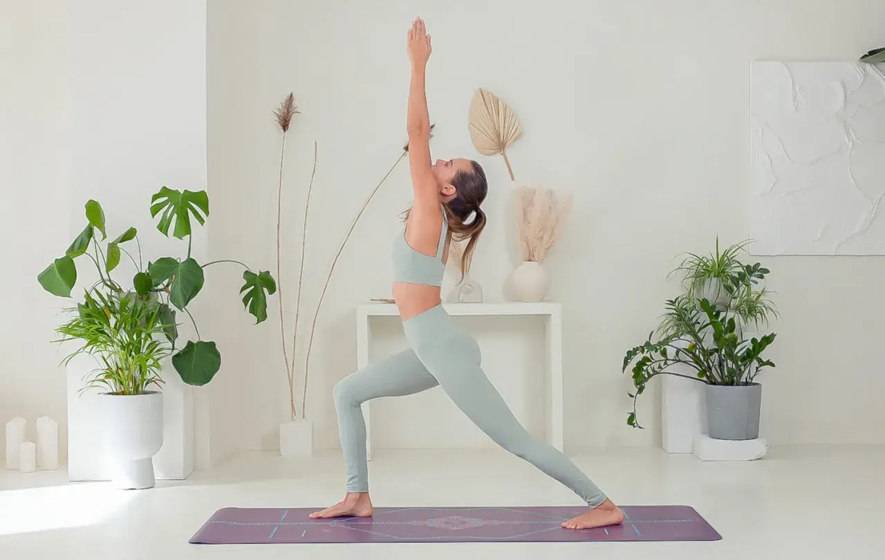 Donna in posa yoga, che mostra i benefici di una pratica regolare. L'ambiente è luminoso e pieno di piante, evocando calma e benessere.