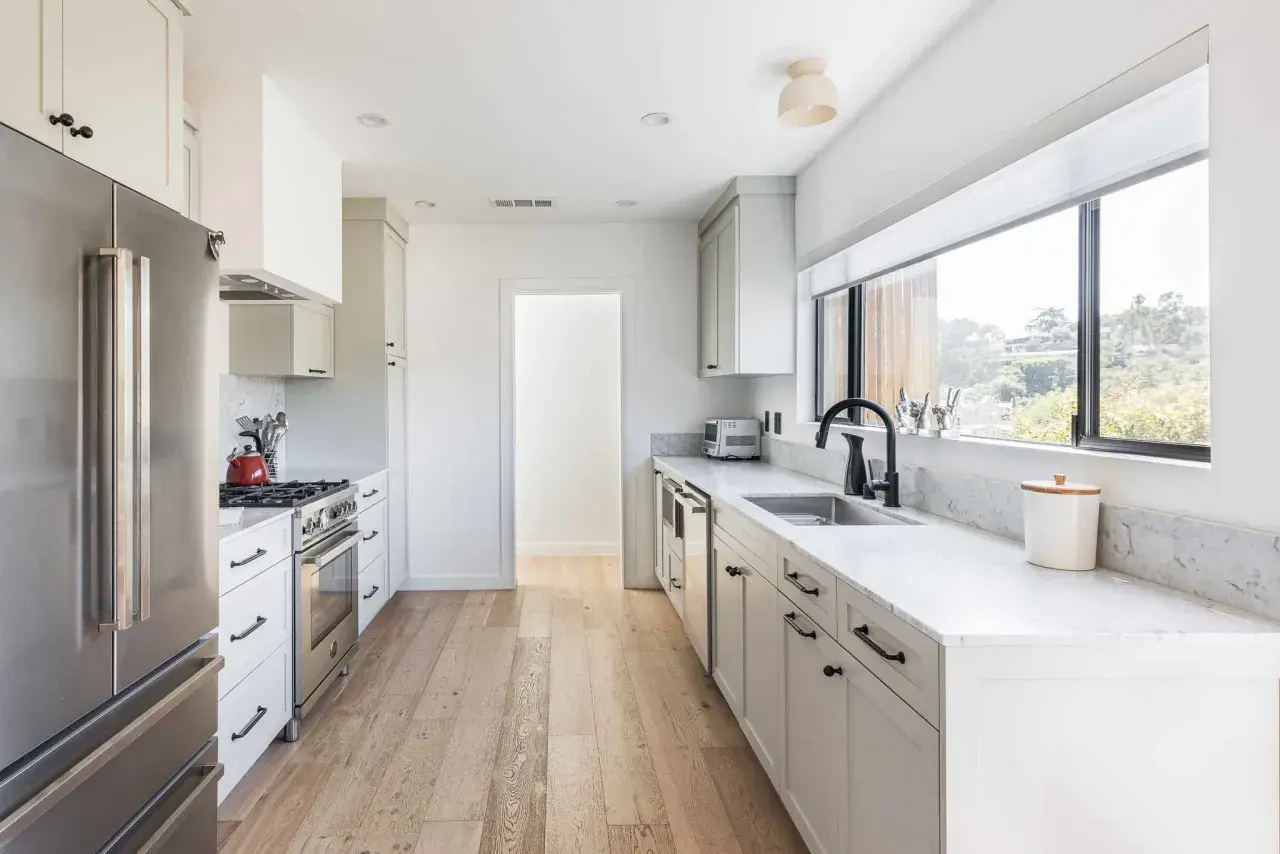 A bright, modern galley kitchen with white cabinets, stainless steel appliances, and a large window overlooking greenery.