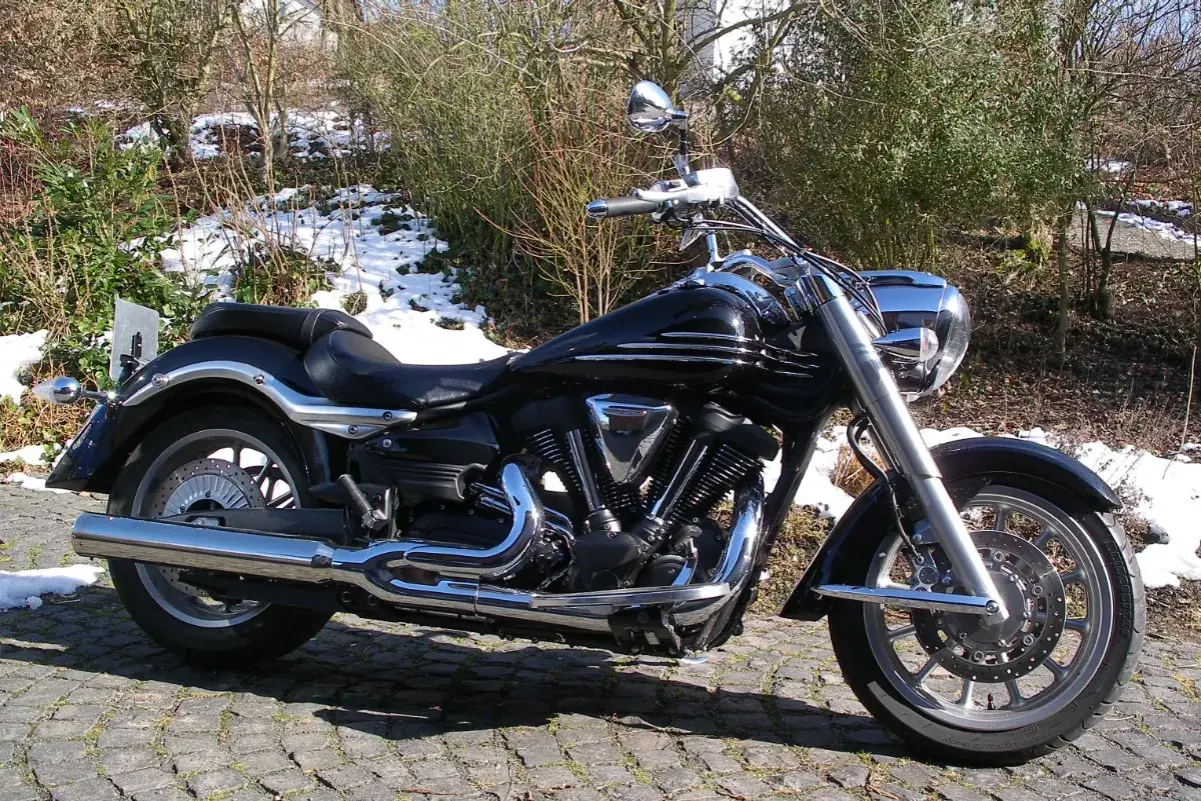Błyszczący czarny motocykl Yamaha Raider 1900, z chromowanymi detalami, zaparkowany na bruku. W tle zima i krzewy.