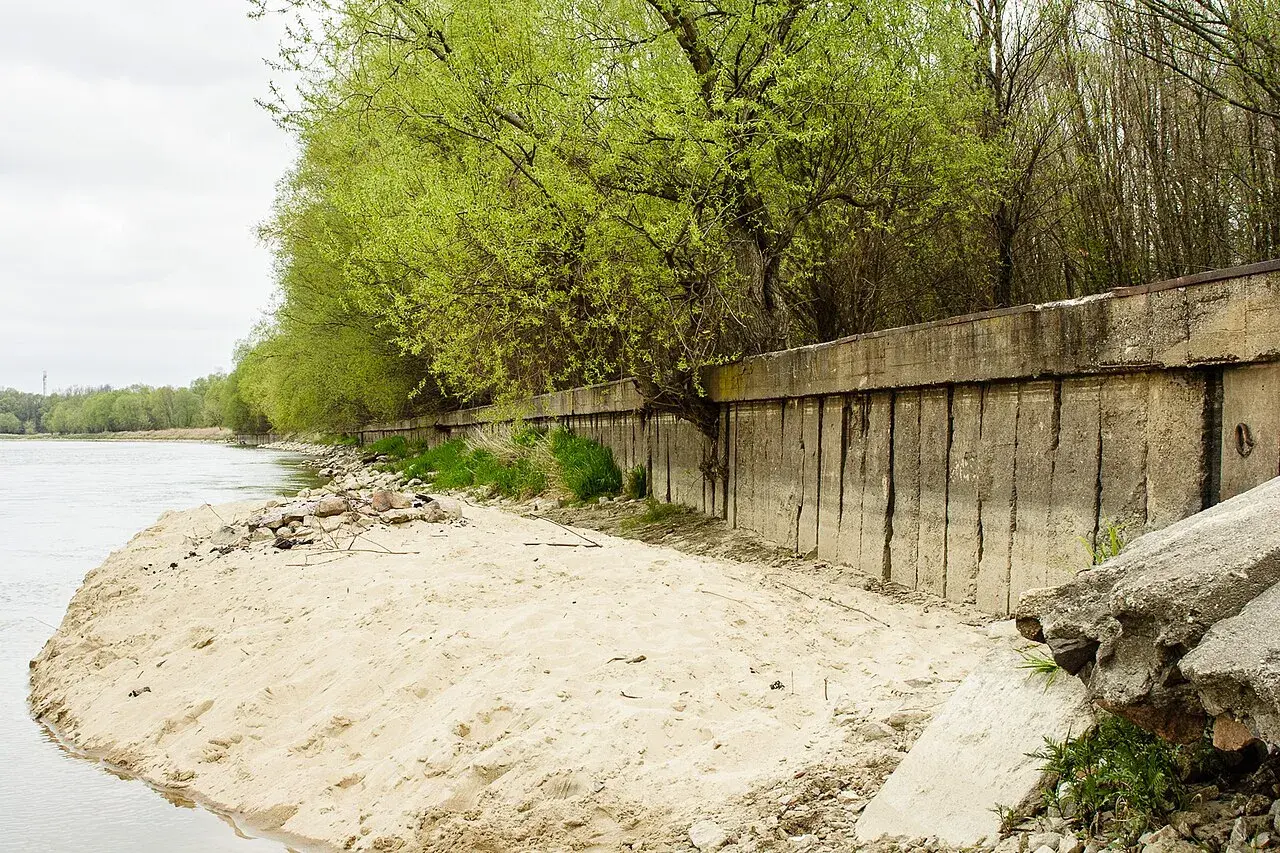 Betonowa opaska na rzece chroni brzeg przed erozją. Wiosenne drzewa tworzą zieloną ścianę nad wodą.