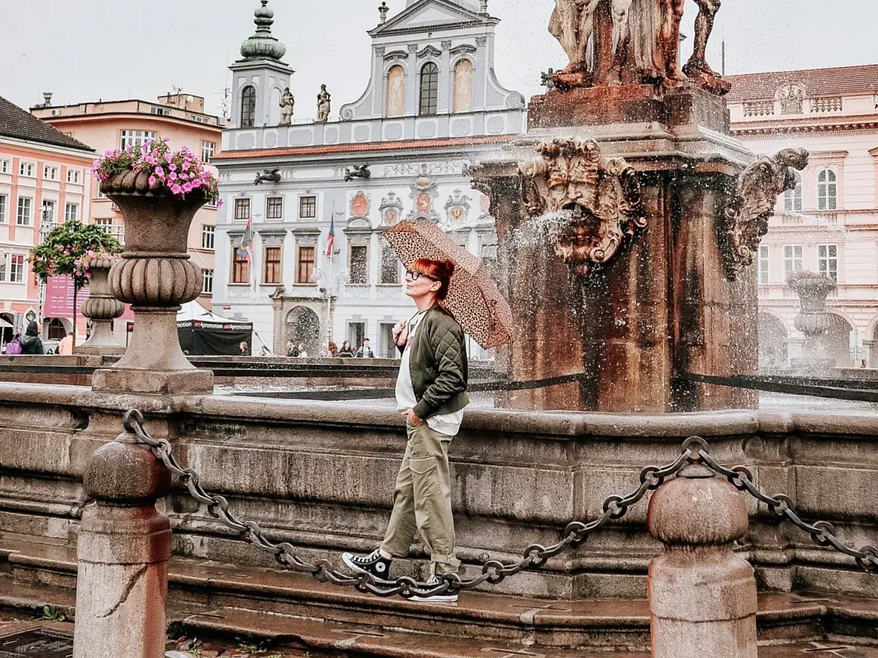 Regenschirm-Schwung am Brunnen in Budweis Stadt. Die Frau genießt die frische Brise, während die historische Kulisse im Hintergrund erstrahlt.