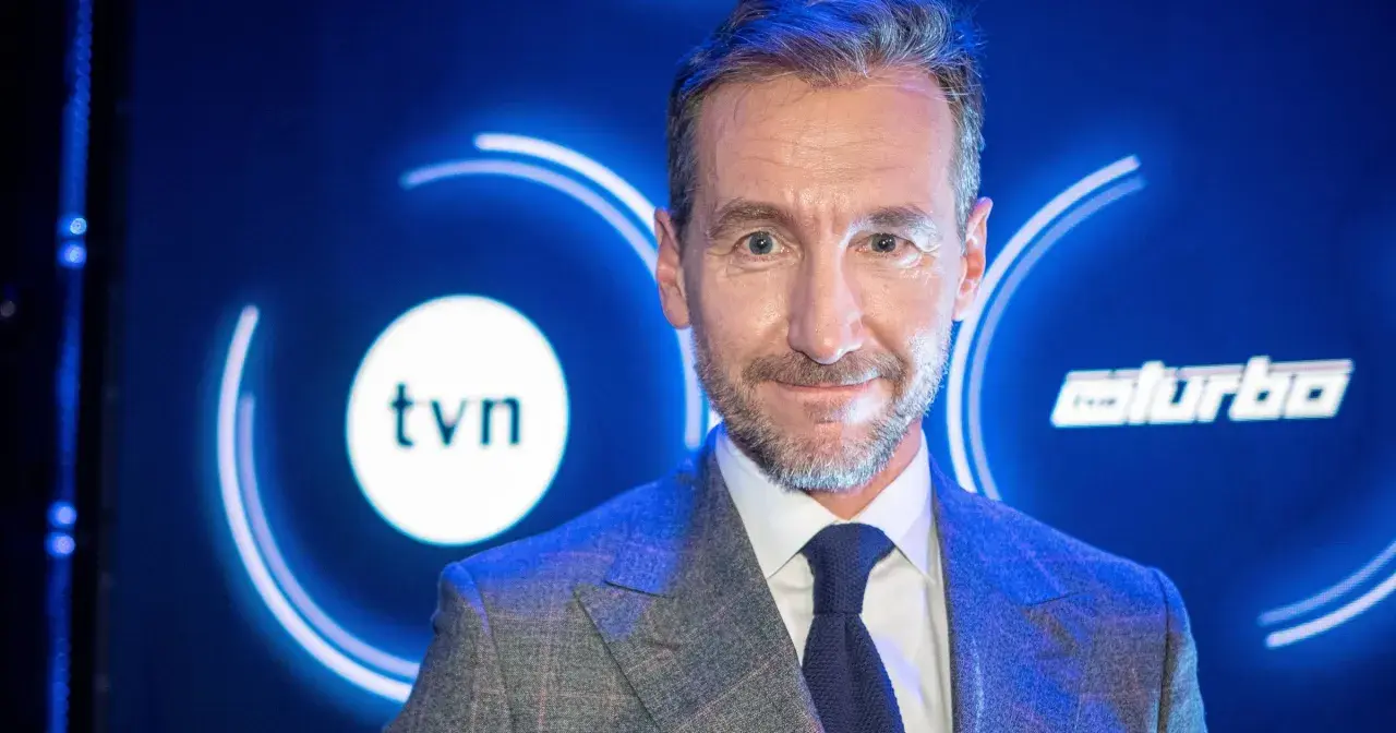 Uśmiechnięty mężczyzna w garniturze na tle logo TVN. Jeden z dziennikarzy Wiadomości TVN, gotowy do przekazania najnowszych informacji.