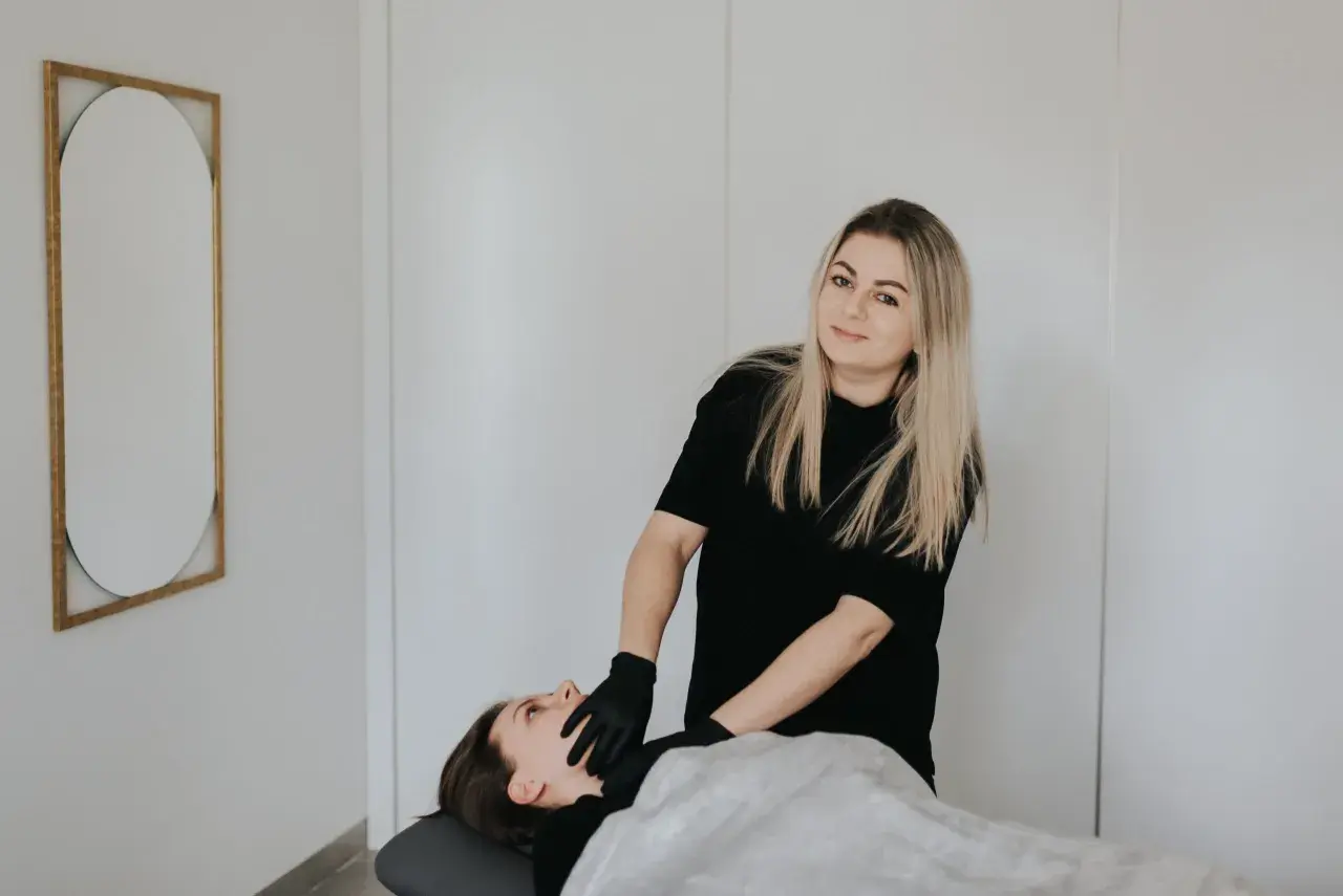Terapeuta w czarnych rękawiczkach wykonuje zabieg rehabilitacji głosu u pacjentki leżącej na kozetce.