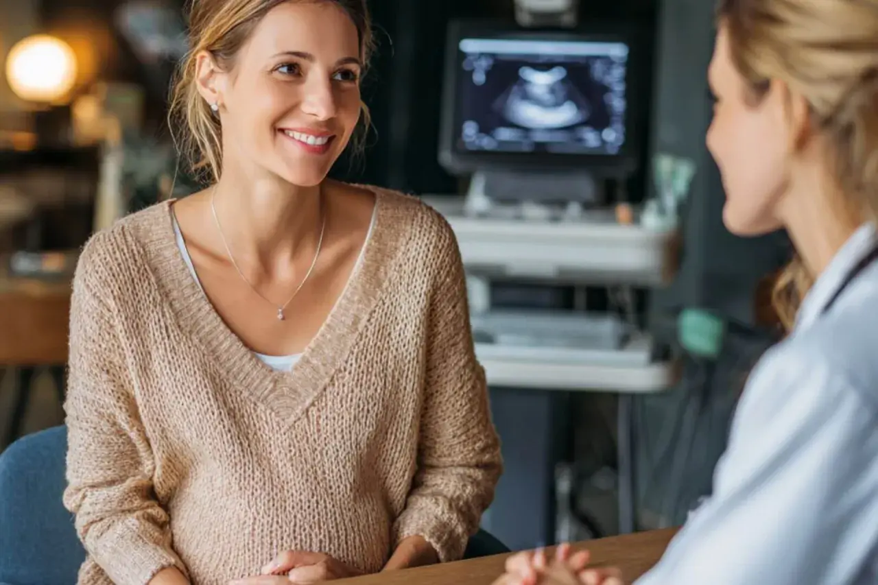 Mujer sonríe mientras habla con su doctora, quien mira una ecografía. Es el momento perfecto para saber cuando decir que estas embarazada.