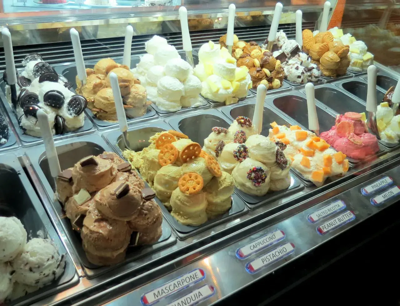 Vetrina piena di gusti di gelato, alcuni con panna montata e decorazioni golose.