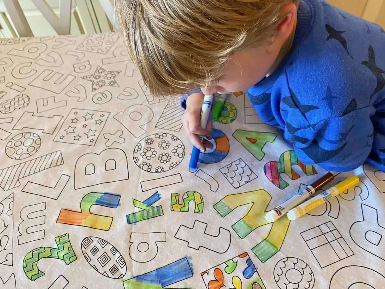 Niño rubio colorea un mantel con letras y dibujos. El **cuadrado para colorear** se llena de vida con sus rotuladores.