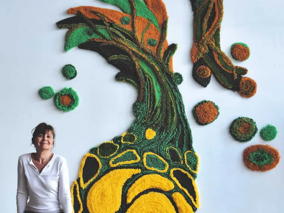Une designer textile pose devant sa création murale en relief, un arbre aux couleurs vives.