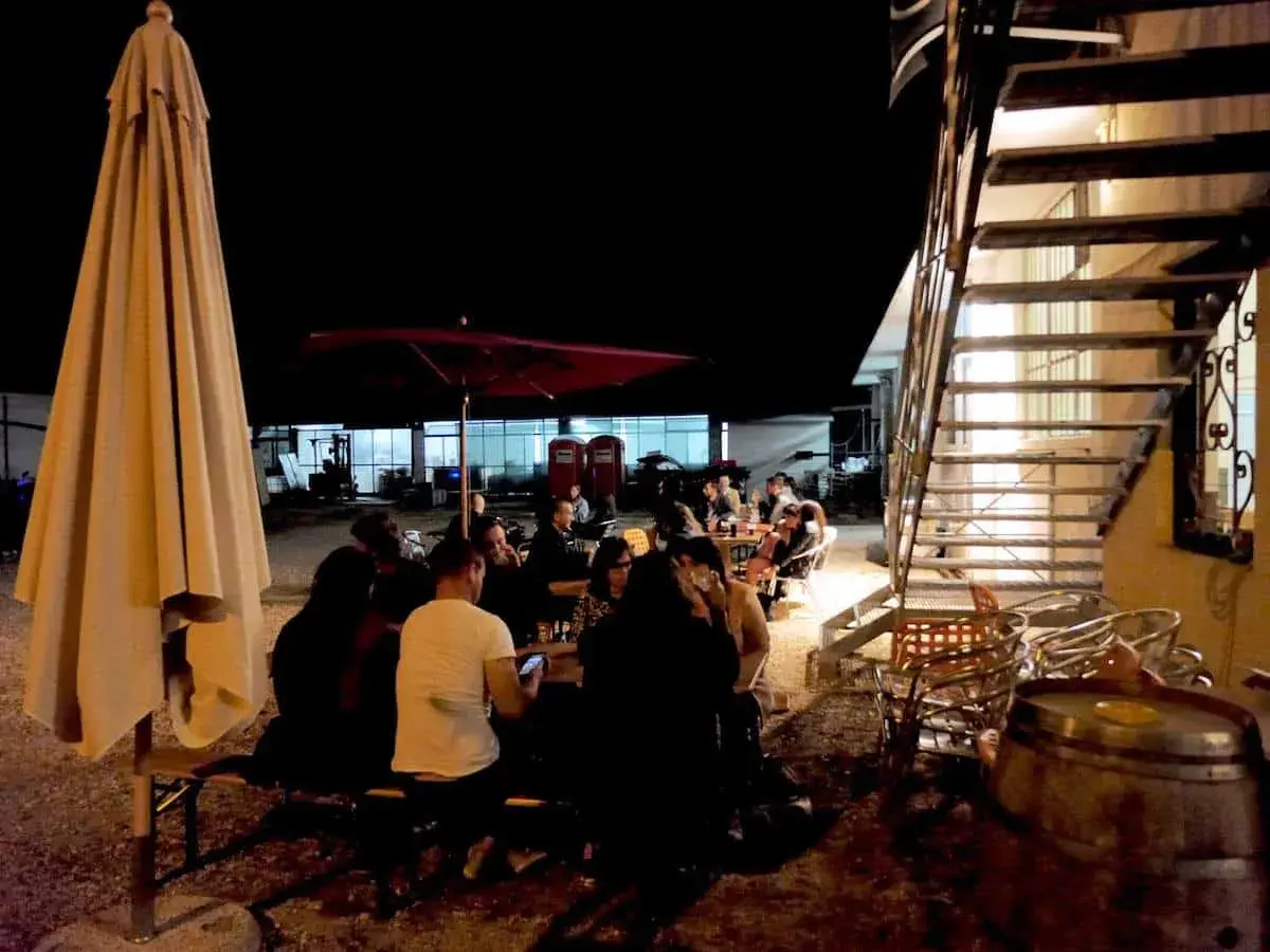 Serata al birrificio Mastro Matto: gente seduta ai tavoli sotto un grande ombrellone, con una scala metallica sullo sfondo.