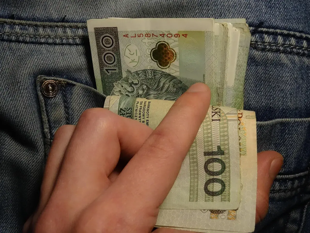 Dłoń wyciąga banknoty 100 zł z kieszeni dżinsów. Może to symbolizować walkę z zatorami płatniczymi lub po prostu gotówkę.