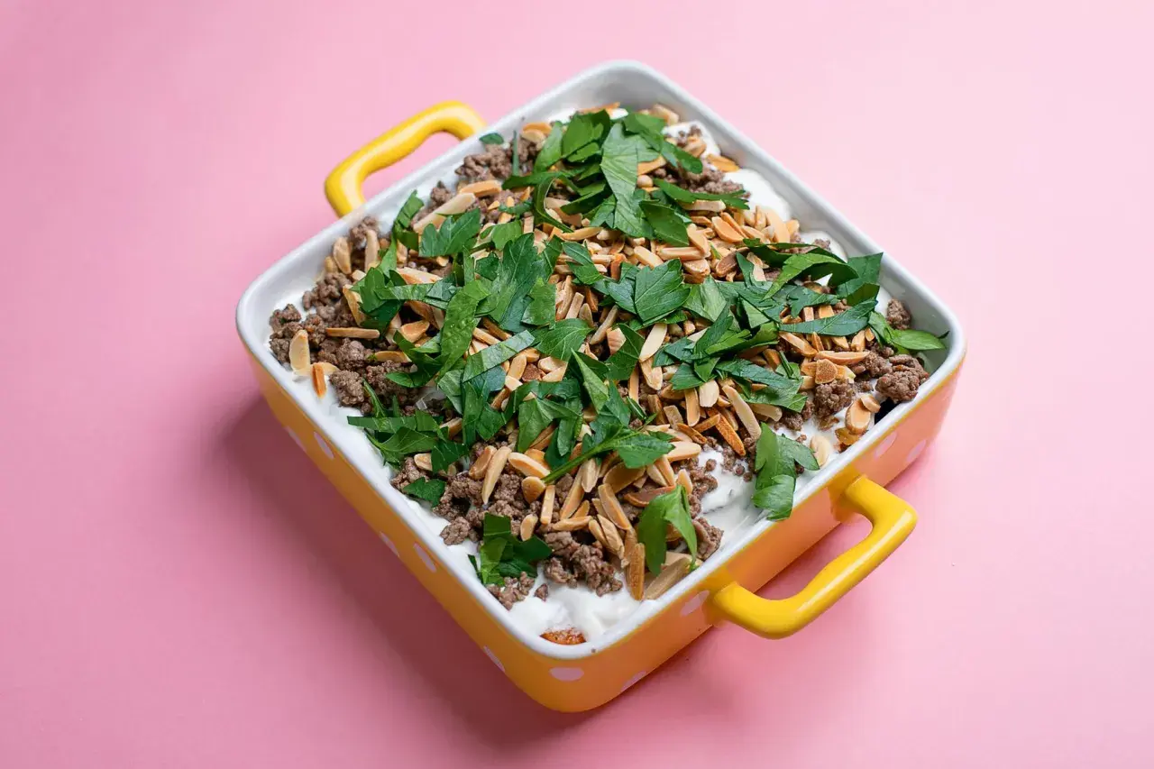 Un plat de fatteh aubergine garni de viande hachée, d'amandes effilées et de persil frais, dans un plat jaune à pois blancs.