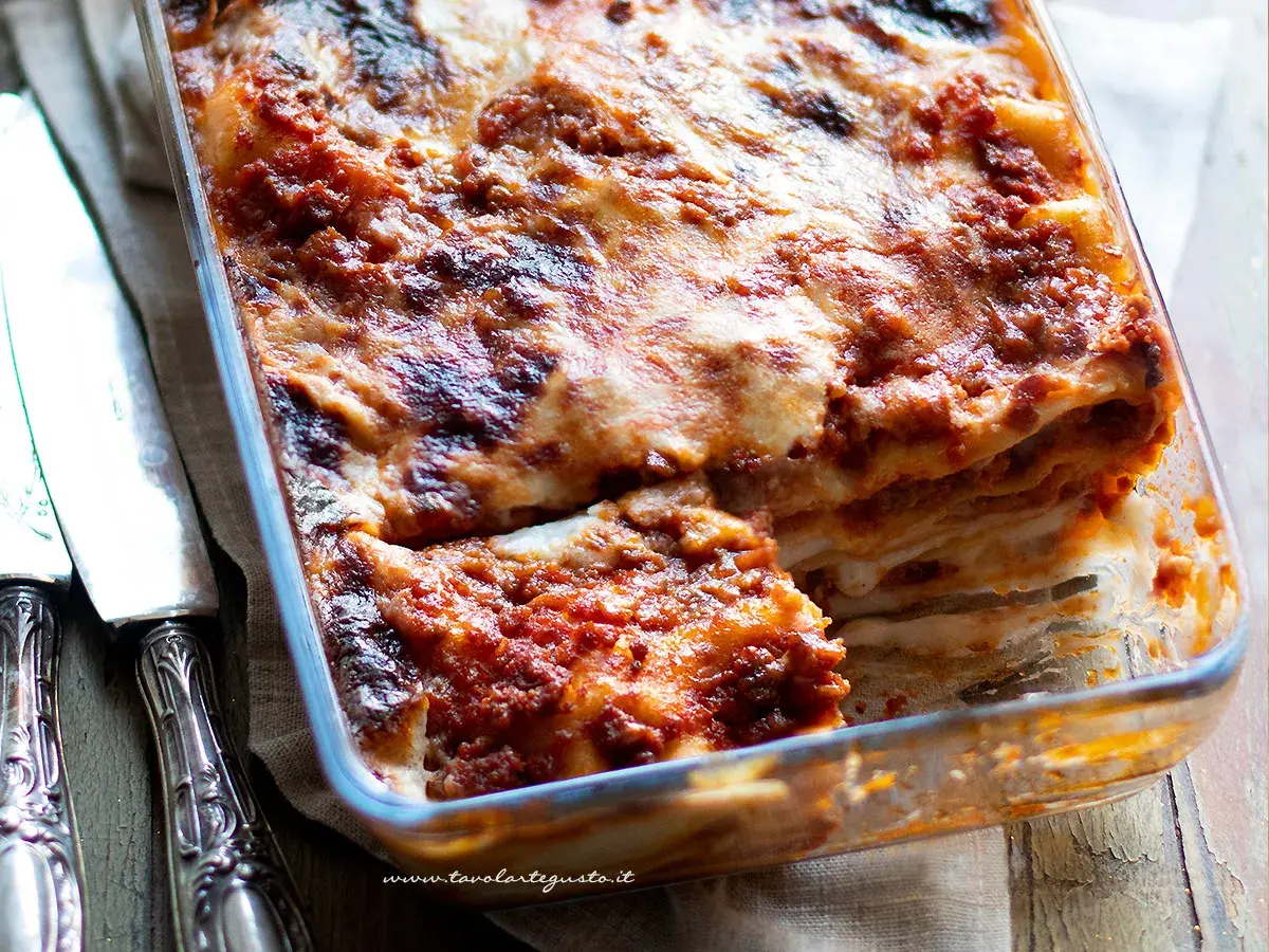 Lasagna al ragù appena sfornata, dorata e filante, in una teglia di vetro, con posate d'argento a lato.