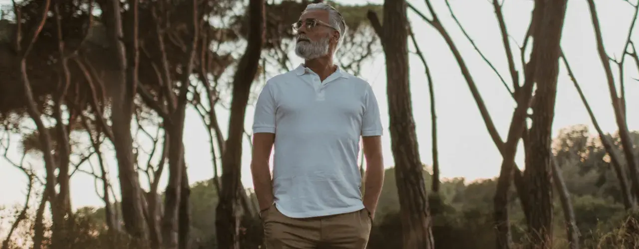 Älterer Mann mit Bart in weißem Polo und brauner Hose, steht im Wald. Die polo farben sind klassisch und zeitlos.