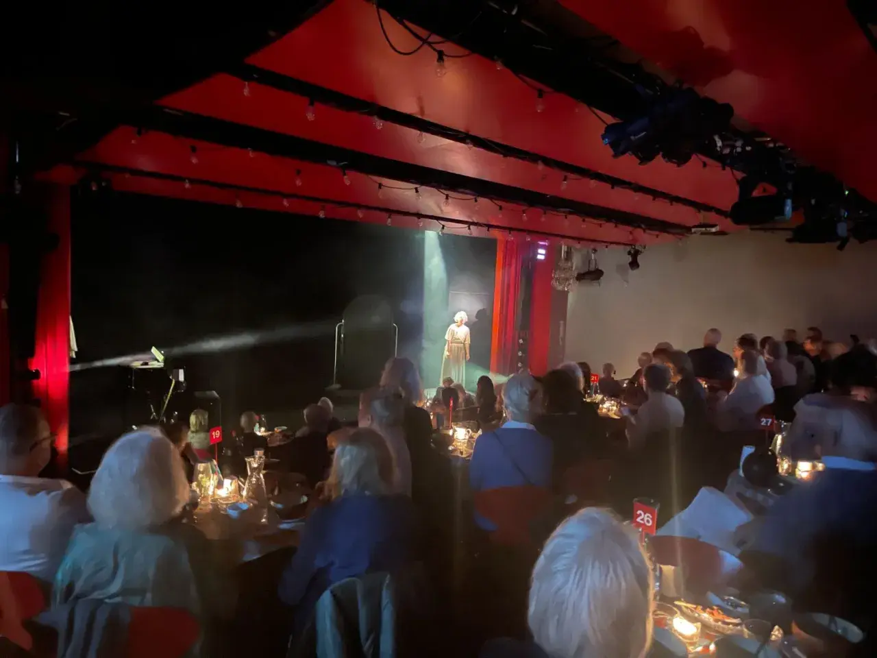 En föreställning på soppteatern Stockholm. En ensam artist står på scenen i strålkastarljus, publiken sitter vid dukade bord.