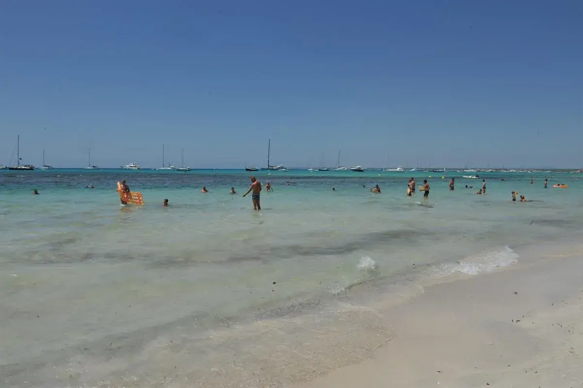 Słoneczny dzień na plaży Es Trenc. Ludzie kąpią się w turkusowej wodzie, a na horyzoncie widać żaglówki.