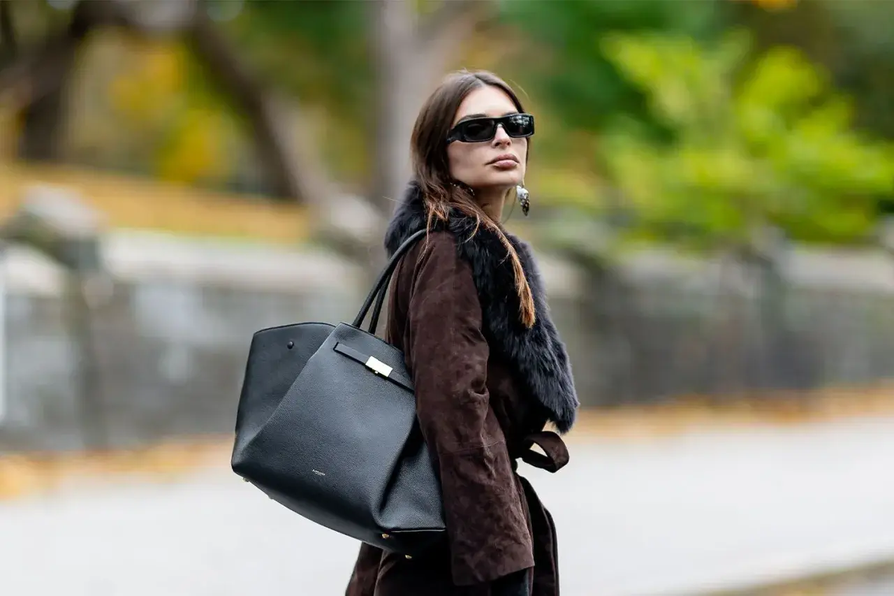 Mujer con abrigo marrón y bolso negro de Demellier, gafas de sol y pendientes llamativos.