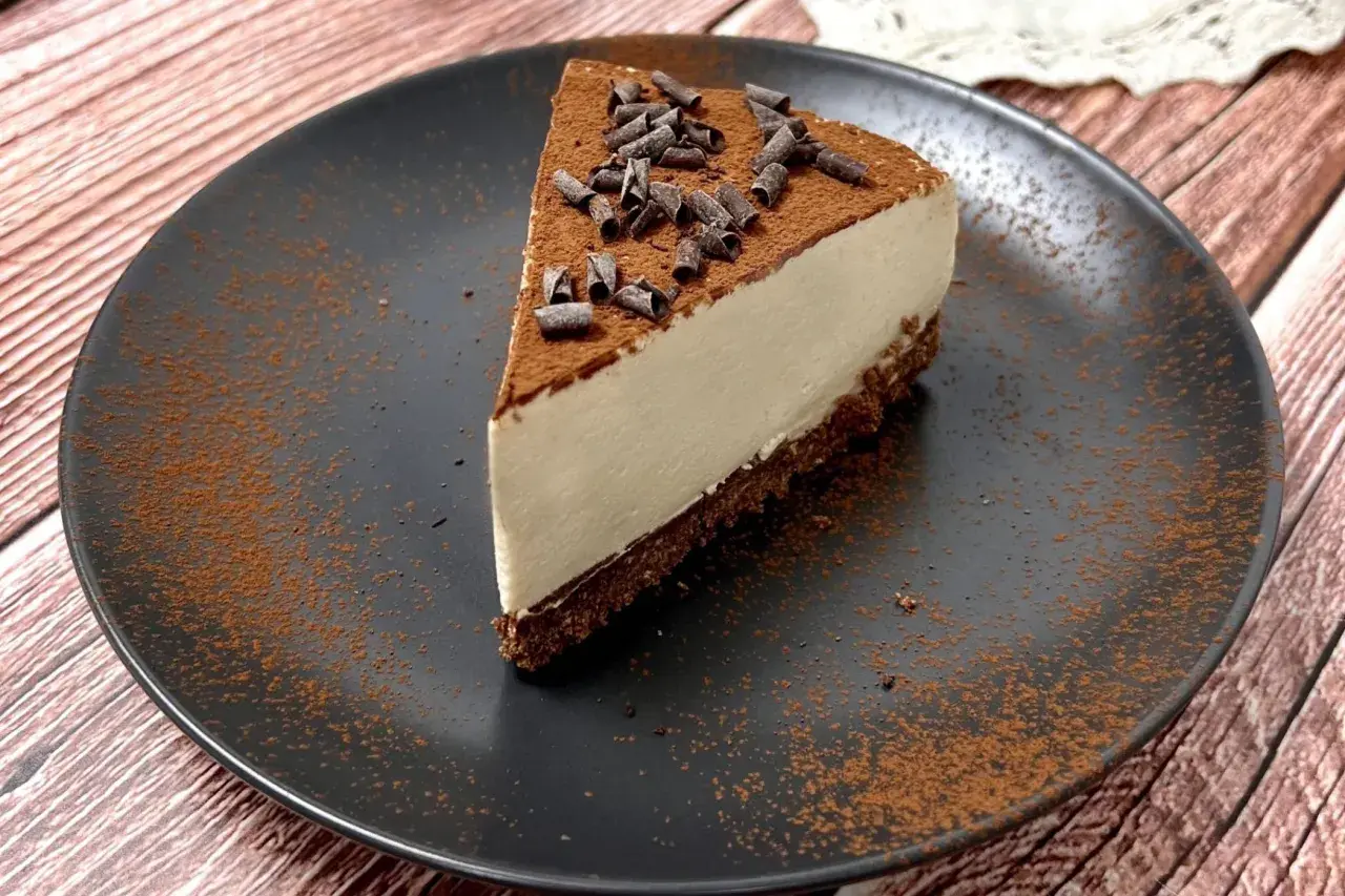 Una fetta di cheesecake al caffè, cosparsa di cacao e riccioli di cioccolato, su un piatto nero.