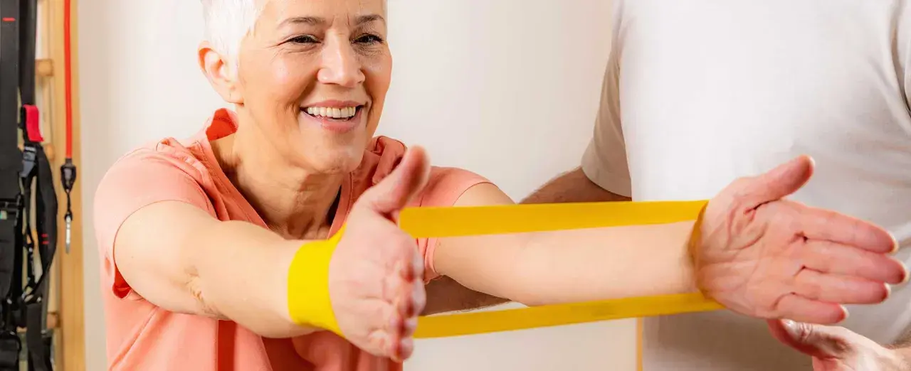 Uśmiechnięta starsza kobieta ćwiczy z fizjoterapeutą, używając żółtej gumy rehabilitacyjnej.