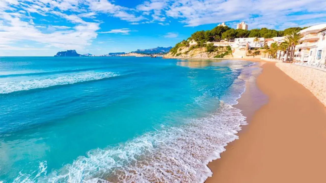 Una playa de arena dorada y aguas turquesas, con edificios blancos y vegetación en la ladera, que evoca las 10 mejores playas de Alicante.