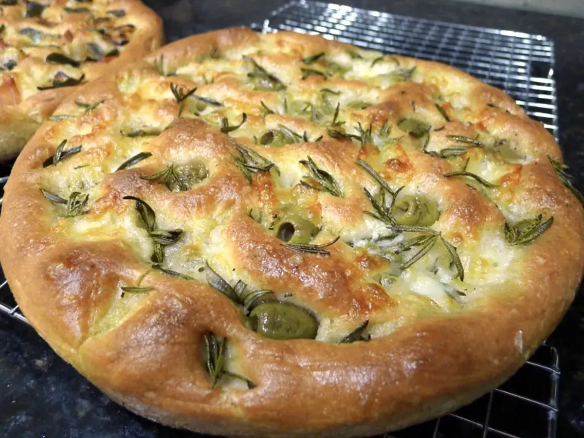 Une focaccia pizza dorée, garnie d'olives vertes et de romarin frais, sort du four.