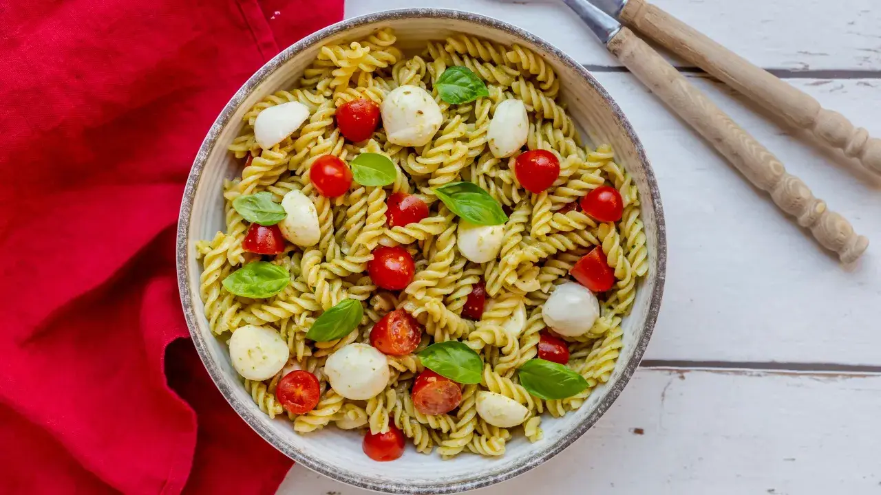 Un piatto invitante di pasta fredda al pesto, arricchito da ciliegini succosi e mozzarelline fresche, guarnito con basilico profumato.