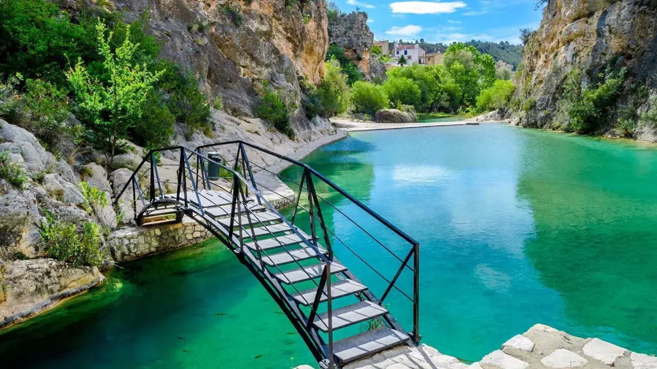 Puente metálico sobre las aguas turquesas del **río Bolbaite**, rodeado de rocas y vegetación exuberante.