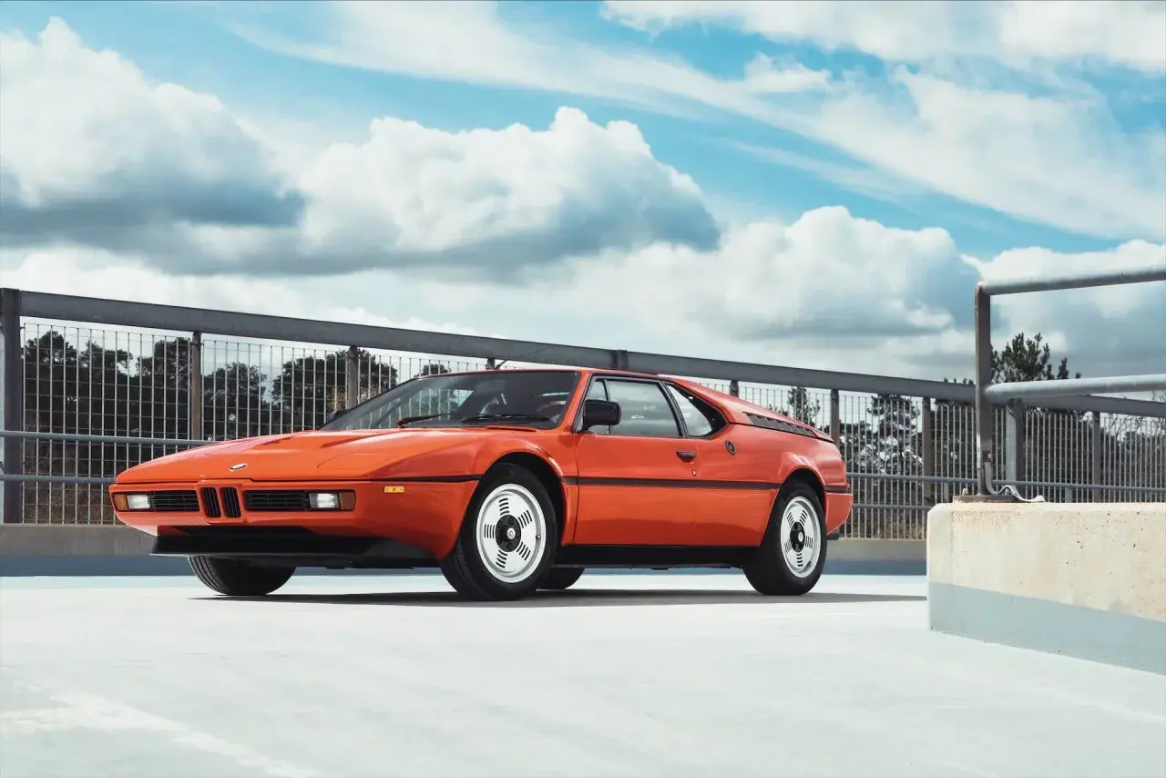 Pomarańczowe BMW E26, ikona lat 80., prezentuje się na tle błękitnego nieba z chmurami.