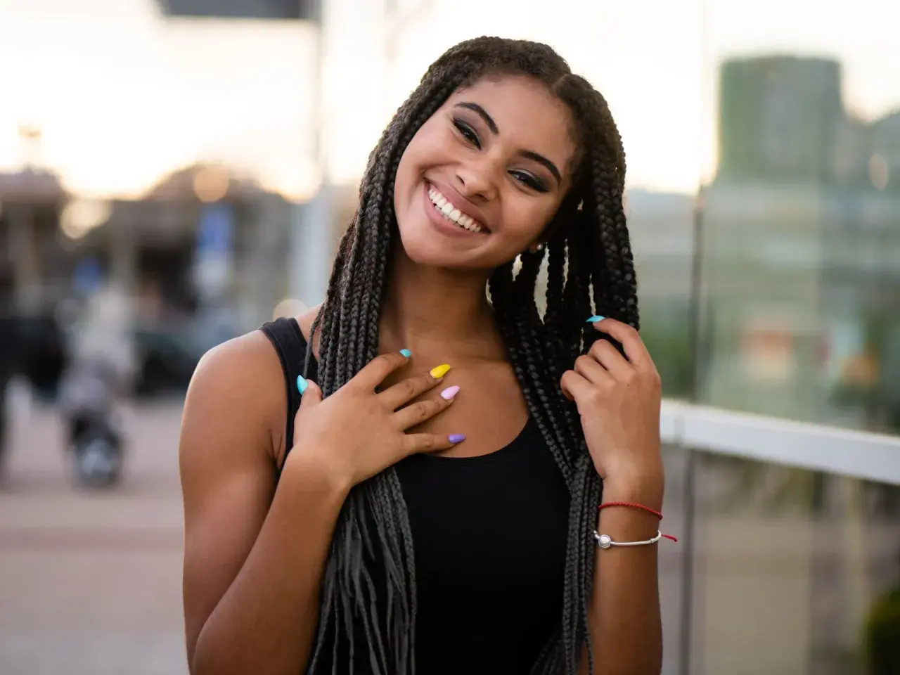 Joven sonriente con trenzas africanas, uñas de colores y un brazalete rojo.